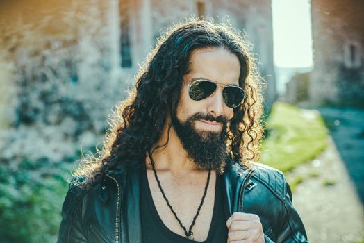Self confident handsome man in sunglasses. Macho man walking outside alone. Stylish young fashionable man with long wavy hair wearing bikers leather jacket and aviator sunglasses