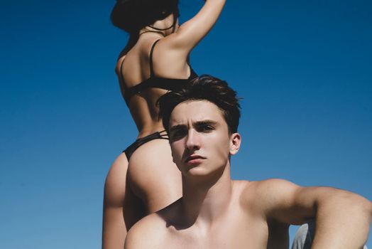 Beautiful young couple of lovers isolated at clear blue sky background. Sexy hot woman with fit sexy ass in bikini standing behind her handsome boyfriend. Sexy desire and love concept