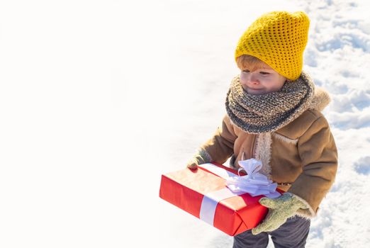 Kid in winter clothes holds Christmas gift. Kid during stroll in a snowy winter park. Beautiful winter nature. Amazing winter park. Adorable kid with cute face. Ptoto with empty copy space