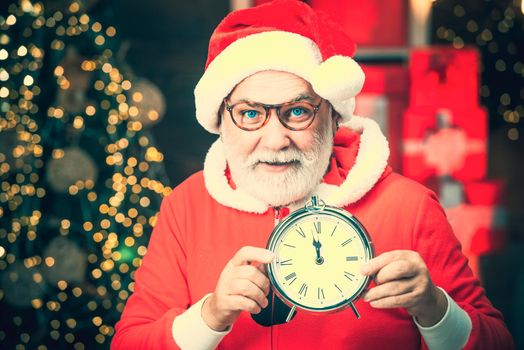 Photo of Santa holding clock showing five minutes to midnight. Cheerful Santa Claus holding alarm clock on fireplace and Christmas Tree background. Greeting Christmas card