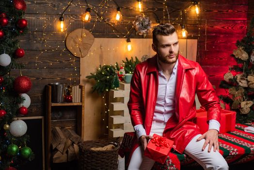 Sexy hot boy wearing red christmas jacket and holding christmas present in his hand. Christmas time. Present box. Shopping sale. Bearded handsome man at full christmas decorated interior