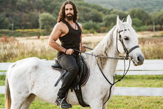 Young jockey training his horse for a ride. Good looking, hunky cowboy rides horse at beautiful country side nature. Equestrian rides his white well looking horse