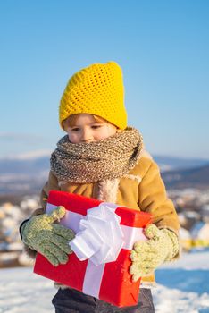 Happy child holding Christmas presents. Christmas holidays. Cute preschooler boy in a knitted yellow hat holding a red box with a gift. Beautiful winter nature. Happy new year and merry christmas