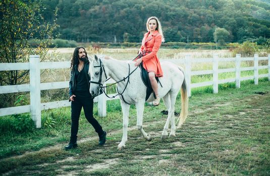 Couple in love walking with horse on countryside. Romantic love story of sensual couple. Woman riding grey arabian horse in pink dress. Handsome bearded man driving horse for a bridle and looking at his girlfriend