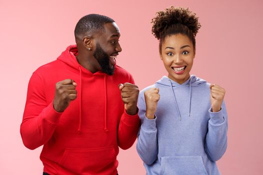 Happy enthusiastic thrilled young african american couple clenching fists joyfully surprised winning awesome honeymoon trip tickets standing pink background triumphing smiling broadly celebrating.