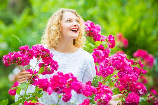 Beautiful woman with spring blooming rose flowers. Young smiling girl enjoys pink rose flowers. Summer time. Happy woman walking at rose garden. Pink rosebush blooming