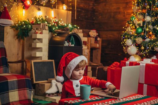 Santa helper baby boy with laptop. Bright New Years interior. Xmas tree. Indoor. Child at Christmas tree and fireplace on Xmas eve. Christmas time. Christmas attributes