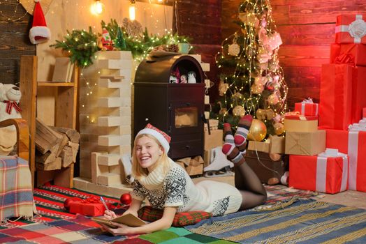 Christmas magic concept. Happy winter holidays at cozy decorated home with family. Teenage blonde girl in knitted sweater dress and red hat writing a wish list of presents to Santa