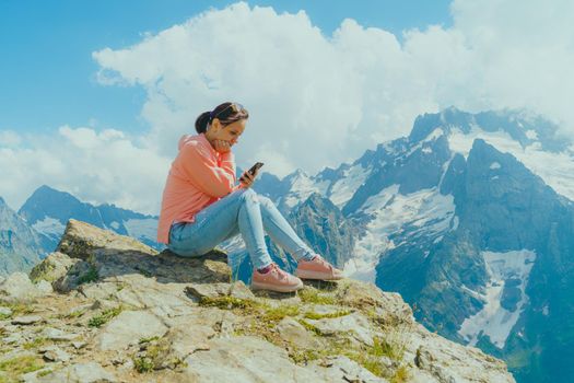 Full body woman sitting on rock and browsing smartphone against cloudy sky on sunny day in mountains