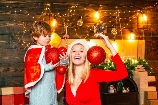 Toddler with mom in the festively decorated room with Christmas tree and decorations. Portrait of happy positive woman sitting on chair against wooden background in the decorated room