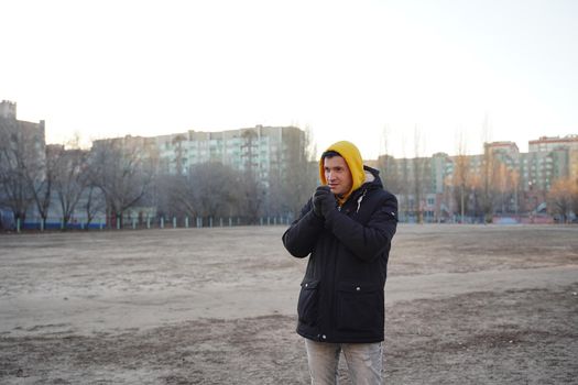 Young frozen man in yellow hood and black jacket. Adult male warming hands in gloves with his breath in cold weather