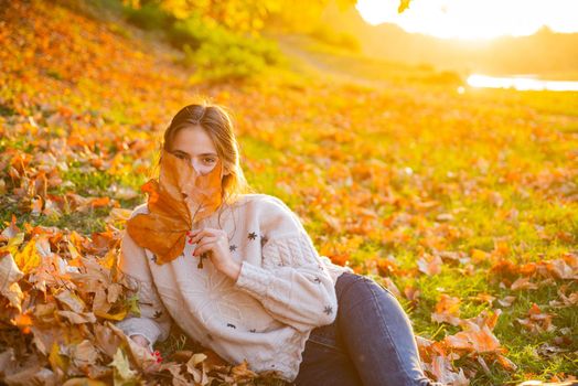 Wonderful brunette woman hide her face with yellow autumnal leaf. Happy beautiful girl have a good time resting in a autumnal golden park. Warm sunny autumn evening