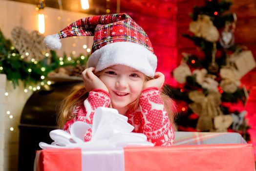 Little cute girl in Santas's hat chherful smile. Christmas gifts. Little child having fun and playing near Christmas tree indoors. New Year interior. Merry Christmas and happy new year