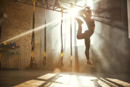 Perfect flexibility. Full length of athletic woman in black sport clothing jumping while exercising in professional semi-dark gym. Sport concept. TRX Training. Gymnastics