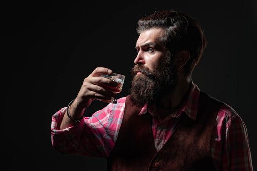 Handsome bearded businessman is drinking expensive whisky. Confident bearded man in black suit with glass of whisky in loft