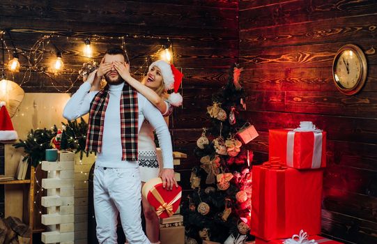 Happy girlfriend prepare christmas surprise for her boyfriend covering his eyes with hands. Present box. Full decorated interior for new year holidays. Celebrating Christmas together concept