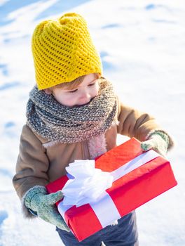 Little boy in warm winter clothes holding gift box and having fun. Bright clothes for children. Beautiful winter nature. Snow-covered forest path. Snowy cold weather concept