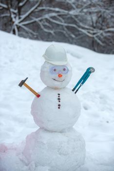Snowman worker with hammer on snow background. Funny snowman in work helmet on snowy field. Handmade snowman in the snow outdoor