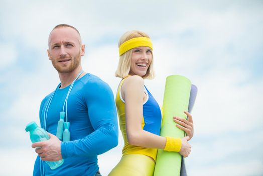 Muscular sport trainer with cheerful smiling young woman holding yoga mat at cloudy sky background. Couple doing sport activities. Young couple in love workout outdoor