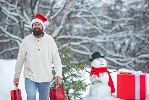 Funny Santa man posing with red gift box on winter weather. Handsome Winter Man with gift and snowman in frosty winter Park