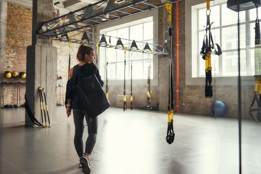 Morning workout. Back view of young athletic woman carrying her sport bag and looking aside while standing at empty gym. Professional sport. TRX Training. Workout