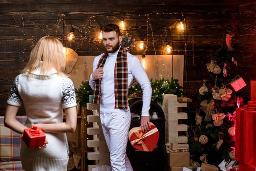 Beautiful girl surprising her boyfriend with a present hidding it behind back. Young couple in love at nicely decorated Christmas interior exchanging Christmas presents. Merry Christmas concept