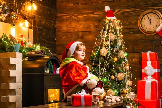 Little Santa Claus helper elf with a magic gift for Christmas. Boy cute child cheerful mood play near christmas tree. Christmas interior. Merry xmas and happy new year