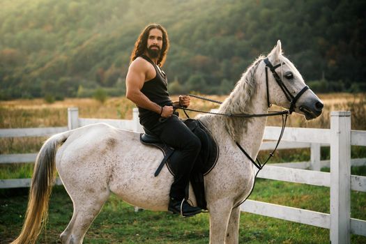 Good looking, hunky cowboy rides horse at beautiful country side nature. Equestrian rides his white well looking horse. Young jockey training his horse for a ride