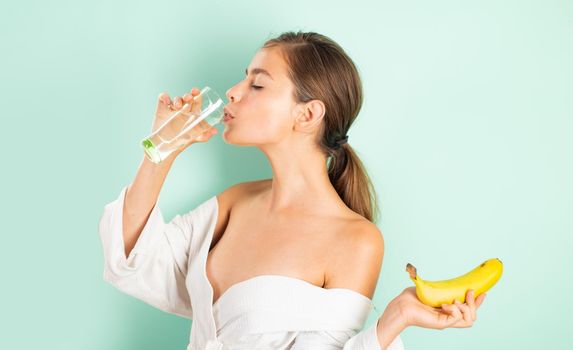 Natural beautiful girl with bare shoulders and clean skin drinks glass of water and holding banana in her hand. Health and beauty concept