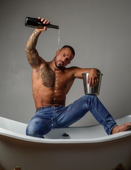 Strong muscular tattoed man holding champagne bottle and posing in bathroom. Handsome bearded shirtless man in jeans with sexy body in bathroom. Sexual macho man in bath