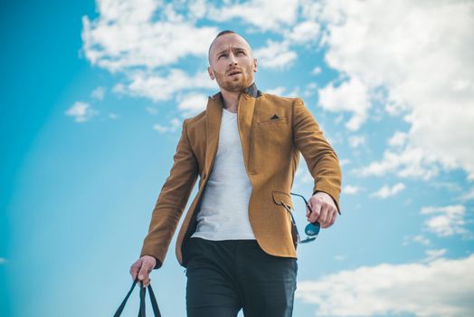 Man with confident face and brutal style. Advertising for clothing store. Standing on the pure blue sky background next to copyspace. Fashionable man in leather jacket