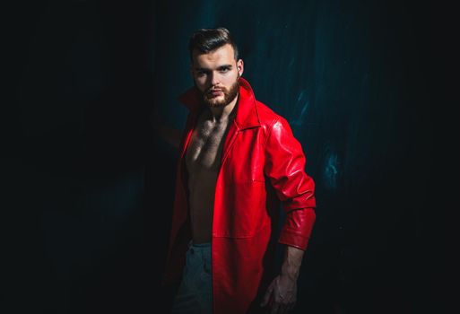 Fashionable man in red jacket is standing on dark background and looking at camera. Portrait of thinking stylish young man. Man with confident face and brutal style