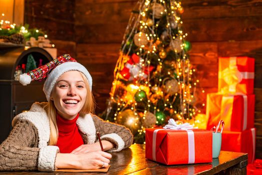 Cheerful smiling blonde woman writing a letter to Santa with her wish list of presents. Christmas presents gifts concept. Joy and happiness. Christmas. Xmas tree