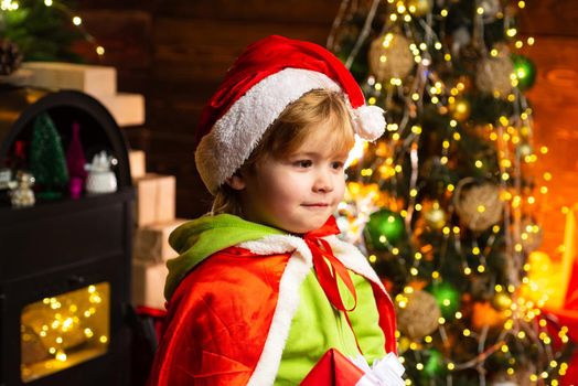 Happy little child in Santa's hat at Christmas tree background. Christmas time. Boy cute child cheerful mood play near christmas tree. Gifts for winter holidays near fire place