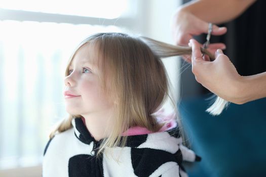 Hairdresser cutting hair of little girl in beauty salon. Hairdressing services for children concept