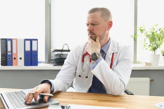 Pensive doctor looking at laptop screen and typing on keyboard in clinic. Online medical training concept