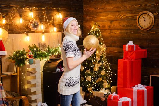 Happy smiling blonde girl wearing Santa's hat with fur and cozy warm sweater with deers and snowflackes. Snow Maiden beautiful girl hold a decorate ball at christmas background