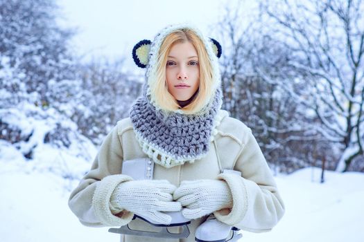 Young blond woman in sweater and funny hat holding white skates over shoulder in freezing winter day. Winter holiday. Girl playing with snow in park