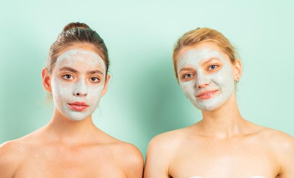 Two natural beautiful friends girls with clay mud mask at face do they healthy skincare. Health and beauty concept. Close up portrait of best friends isolated at pale blue background