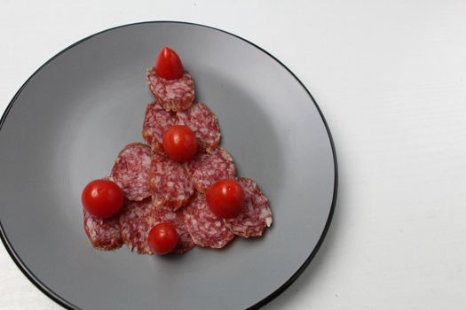 edible Christmas tree made of sausage sausage and cherry tomato lies on a gray plate on a white light background with space for text.