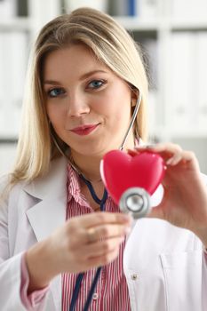 Beautiful smiling blond female doctor hold in arms red toy heart closeup. Cardio therapeutist student education CPR 911 life save physician make cardiac physical pulse rate measure arrhythmia