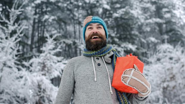 Hipster santa in cold snowy winter forest. Happy holiday and xmas. Bearded man in santa hat at new year. Christmas happy man with beard hold present box.