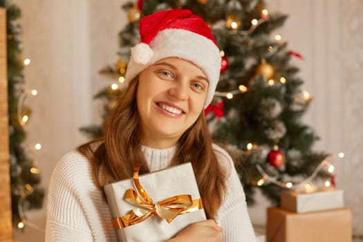 Portrait of smiling woman holding New Year gift in box with gold ribbon, happy female wearing santa hat and white sweater posing indoor with Christmas tree on background.