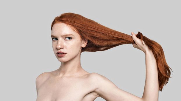 Studio shot of young and confident redhead woman holding her long silky hair while standing against grey background. Beauty concept. Hair care