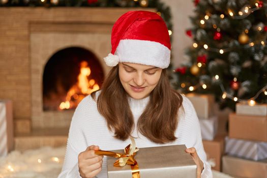 Adorable charming cheerful woman holds gift box and guess what inside, keeping fingers on ribbon and looking on box, sitting with fireplace and Christmas tree on background.