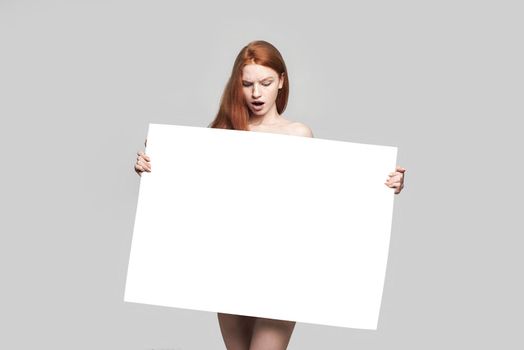 Studio shot of shocked young redhead woman holding empty blank board and looking at it while standing against grey background. Advertising