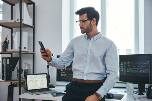 Stock exchange. Concentrated young trader or businessman in formal wear and eyeglasses is looking at trading reports on his smartphone for investment purpose while working at his office. . Crypto currencies. Business concept. Investment concept