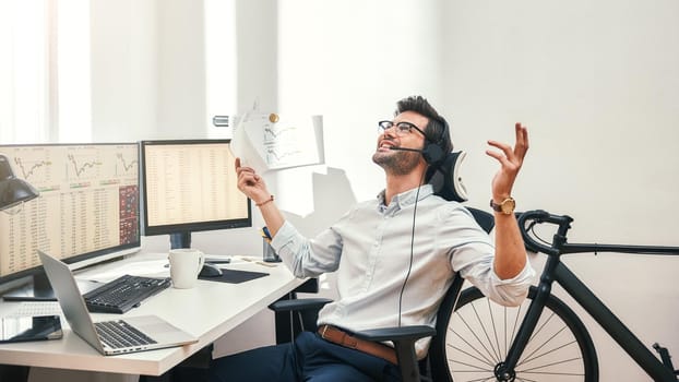 Great Happy and successful bearded trader in headset holding financial report, talking with client and gesturing while sitting in front of monitor screens in the office. Success concept. Trade concept. Call center