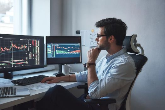 Busy working day. Side view of successful trader or businessman in formal wear and eyeglasses working with charts and market reports on computer screens in his modern office. Stock broker. Forex market. Trade concept