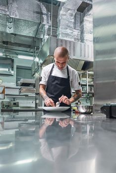 Working in a restaurant. Vertical portrait of professional male chef with tattoos on his arms garnishing his dish on the white plate while standing in a kitchen